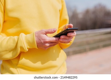 A Man In A Yellow Hoodie Uses A Smartphone. Hands Type On The Phone. A Person Makes An Order Of Delivery Via The Internet. Surfing The Internet On A Mobile Phone. Using Apps, Ordering Taxis And Food