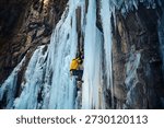 Man in yellow jacket is climbing ice wall at big frozen waterfall with ice axe and crampons against blue ski in the winter mountains. Sport mountaineering and alpinism concept