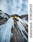 Man in yellow jacket is climbing ice wall at big frozen waterfall with ice axe and crampons against blue ski in the winter mountains. Sport mountaineering and alpinism concept