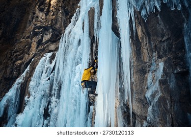 Man in yellow jacket is climbing ice wall at big frozen waterfall with ice axe and crampons against blue ski in the winter mountains. Sport mountaineering and alpinism concept