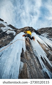 Man in yellow jacket is climbing ice wall at big frozen waterfall with ice axe and crampons against blue ski in the winter mountains. Sport mountaineering and alpinism concept