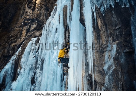 Man in yellow jacket is climbing ice wall at big frozen waterfall with ice axe and crampons against blue ski in the winter mountains. Sport mountaineering and alpinism concept