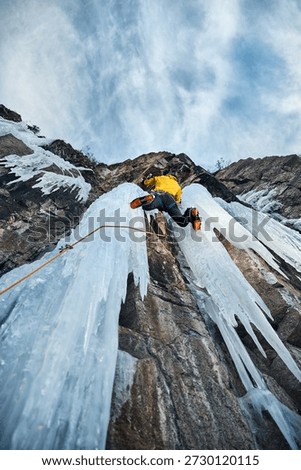 Man in yellow jacket is climbing ice wall at big frozen waterfall with ice axe and crampons against blue ski in the winter mountains. Sport mountaineering and alpinism concept