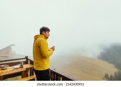 A man in a yellow jacket spends time at a dacha in the mountains, standing on the terrace with a cup in his hands and looking at the misty views of the forest and meadows