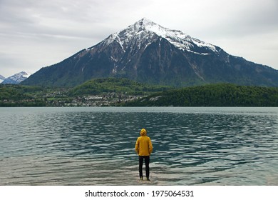 Man with yellow rain jacket in front of huge snow-covered mountain in Switzerland. Mount Niesen with green trees and water of lake Thun.