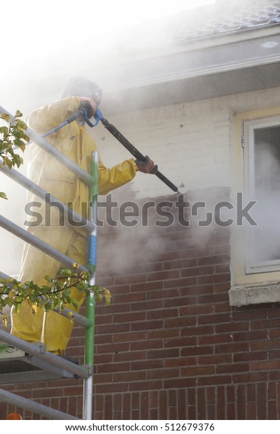 man in\
yellow rain suit cleans paint from brick wall of house facade with\
pressure washer while standing on\
scaffolding