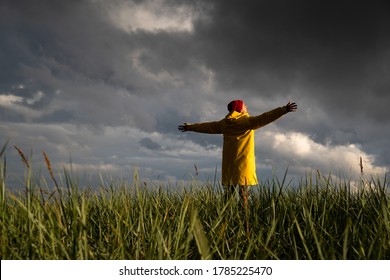 Man in yellow raincoat and red hat with spreading hands wide open standing on the field in rainy weather, looks at dramatic cloudy sky. Autumn season. Enjoying the freedom