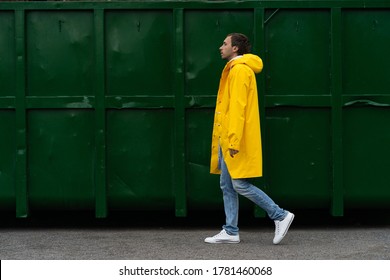 Man in a yellow raincoat walks down the street in the rain weather next to green container, side view. Outdoor.  - Powered by Shutterstock