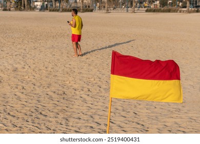 A man in a yellow shirt and red shorts walks across a sandy beach, away from a red and yellow Life Guard Beach Flag planted in the sand.The man seems to be focused on his phone, unaware of the flag. - Powered by Shutterstock