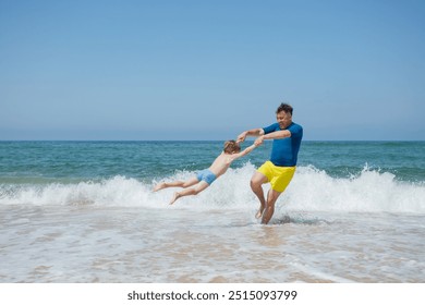 A man in yellow shorts and a blue shirt is playing with a young boy on a beach, spinning him around, the ocean waves in the background - Powered by Shutterstock