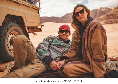 Man and younger woman wearing traditional Bedouin warm coat - bisht -  sitting or laying on ground blanket near off road vehicle back, blurred Wadi Rum desert background - Powered by Shutterstock