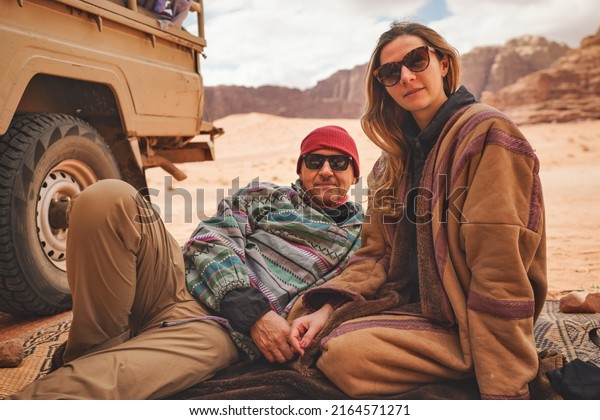 Man and younger woman\
wearing traditional Bedouin warm coat - bisht -  sitting or laying\
on ground blanket near off road vehicle back, blurred Wadi Rum\
desert background