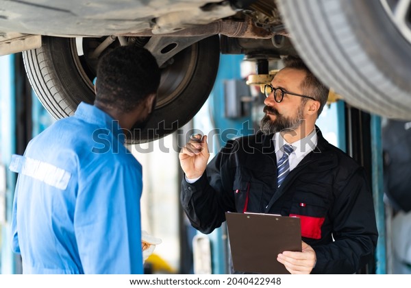 Manager with beard and\
checklist clipboard talk to Expertise mechanic working Under a\
Vehicle in a Car Service station. Car care and automobile service\
garage concept.