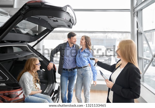 Manager of car dealership\
working with clients, holding folder and giving car keys to buyers\
of vehicle. Couple standing, posing, daughter sitting in car trunk,\
smiling.