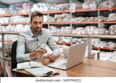 Manager checking his cellphone and using a laptop while sitting at his desk in a carpet warehouse with shelves of stock in the background 