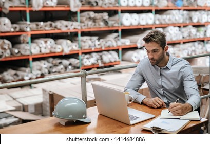 Manager checking inventory and online orders on a laptop while sitting at a desk in a carpet warehouse with shelves of stock in the background