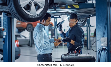 Manager Checks Data On A Tablet Computer And Explains The Breakdown To A Mechanic. Car Service Employees Inspect The Bottom Of The Car With A LED Lamp. Modern Clean Workshop.