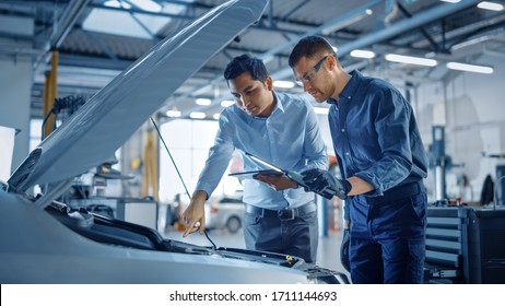 Manager Checks Diagnostics On A Tablet Computer And Explains The Engine Breakdown To A Mechanic. Car Service Employees Inspect Car's Engine Bay With A LED Lamp. Modern Clean Workshop.