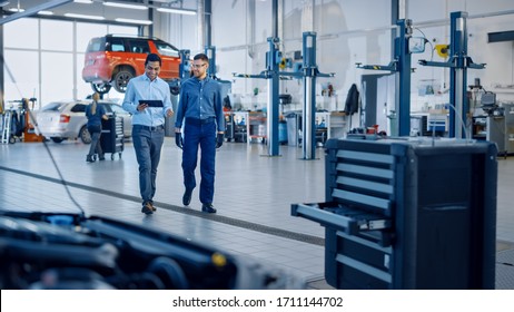 Manager Checks Diagnostics Results On A Tablet Computer And Explains A Vehicle Breakdown To A Handsome Mechanic. Car Service Employees Talk While Walking In A Garage. Modern Clean Workshop.