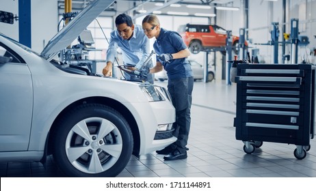 Manager Checks Diagnostics Results On A Tablet Computer And Explains An Engine Breakdown To An Female Mechanic. Car Service Employees Inspect Car's Engine Bay With A LED Lamp. Modern Clean Workshop.