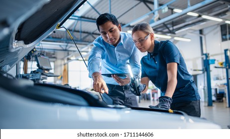 Manager Checks Tasts on a Tablet Computer and Explains an Engine Breakdown to an Female Mechanic. Car Service Employees Inspect Car's Engine Bay with a LED Lamp. Modern Clean Workshop.