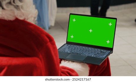 Manager in Christmas adorn shopping mall dressed as Santa Claus holding green screen laptop, looking at clothing store ecommerce website, inputting promotional holiday offers - Powered by Shutterstock