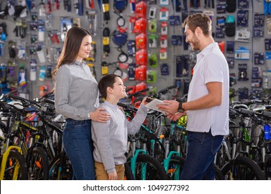 Manager With Digital Tablet Helping To Choose Bicycles To Customers In Bike Shop