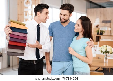 The Manager Of The Furniture Store Holds Samples Of Fabrics For Furniture Upholstery. Near Him Are A Guy And A Girl - Buyers Of Furniture.