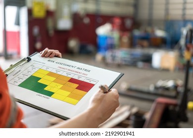 A Manager Is Holding A Pen And Risk Matrix Form With Factory Workshop Place As Blurred Background. Industrial Risk Assessment And Safety Audit Concept Scene. Close-up And Selective Focus.