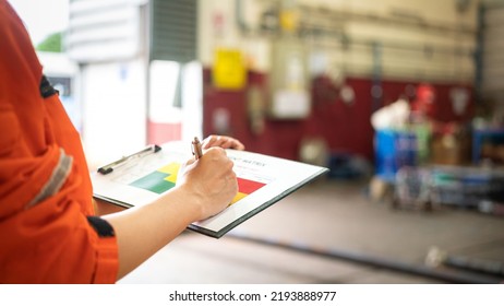 A Manager Is Holding A Pen And Risk Matrix Form With Factory Workshop Place As Blurred Background. Industrial Risk Assessment And Safety Audit Concept Scene. Close-up And Selective Focus At Hand Part.