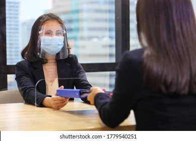 Manager From HR Department Wearing Facial Mask Is Interviewing New Applicant Who Is Handing Her Resume And Profile Through The Partition For Social Distancing, Transaction And New Normal Policy