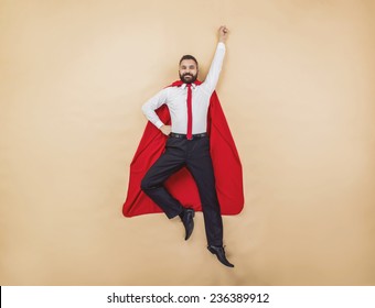 Manager In A Superman Pose Wearing A Red Cloak. Studio Shot On A Beige Background.