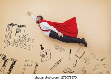 Manager In A Superman Pose Wearing A Red Cloak. Studio Shot On A Beige Background.