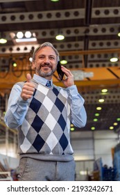 Manager Talking On Smartphone, Looking At Camera And Giving Thumb Up In A Factory. Waist Up Portrait Of Confident, Handsome, Gray-Haired Businessman Or Engineer In Industrial Interior.