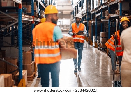 Manager using digital tablet walking through warehouse while workers carrying cardboard boxes, organizing goods distribution and working together in large warehouse