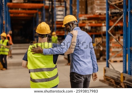 Manager and worker walking together through warehouse, arms around each other's shoulders, demonstrating camaraderie, support, and teamwork in industrial workplace