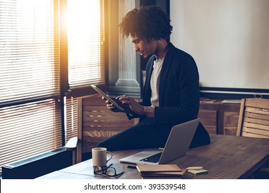 Managing his timetable. Side view of young handsome African man using his digital tablet while sitting on table at his working place - Powered by Shutterstock