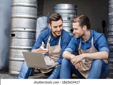 Managing modern brewery, working in warehouse and startup. Smiling young men in aprons sit, look at laptop at data and talking about business in interior of warehouse with metal kegs, free space - Powered by Shutterstock