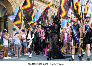 Manchester UK, August 28, 2019. Pride Parade Passing In Front Of Radisson Blue Hotel. Mr Rubber UK And Woman Dressed In Black Lace With Medusa Hair. Black Yellow And Orange Rubber Pride Flag.