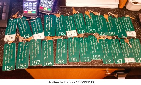 Manchester, United Kingdom - October 23, 2019: Set Of Many Keys On A Busy Reception Desk In A Hotel, With Green Big Tags And Instruction On It To Leave Key At Departure Time. 