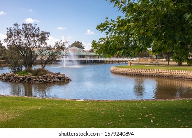 Mandurah, WA, Australia - November 25, 2009: Small island with trees, fountain in blue water of channel waterway and park in front of beige building of The Boardwalk shopping mall under light blue sky