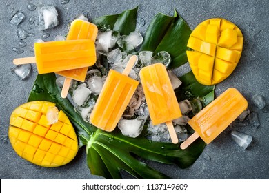 Mango popsicles over green tropical palm leaf on grey stone background. Overhead, top view