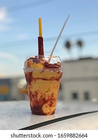 A Mangonada Mexican Frozen Drink, Consisting Of Mango Sorbet, Mango Chunks, Chamoy, Lime, Tajin Powder, And Tamarind Candy, Is Shown Served In A Clear Plastic Cup In A Vertical View.