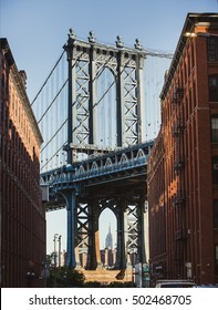 Manhattan bridge enclosed by two brick buildings on a sunny day with Empire State building on background.