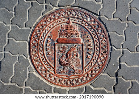 Manhole cover in Kazan, Tatarstan, Russia. Sewer lid with coat of arms of Kazan on road. Top view of metal manhole cap on city street. Traditional dragon emblem and word Kazan on old drain plate.