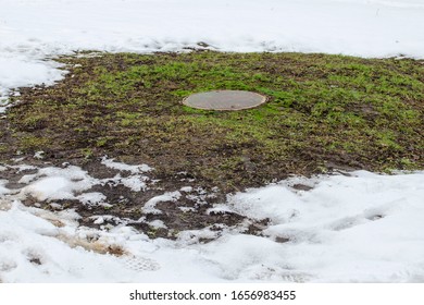 Manhole Of A Sewer Well On A Lawn With Grass Sprouted Around It In Winter Snowy Time