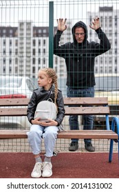 Maniac guy watches through fence at little girl sitting on playground alone, child abuse concept. Caucasian pedophile guy in black wear want to commit crime, look at girl with interest, focus on girl