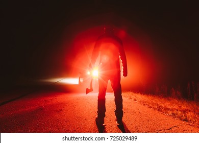 Maniac Killer Near The Car At Night. Silhouette Of A Man With An Ax In His Hand At Night In A Fog