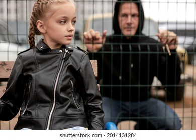 Maniac man watches through fence at little girl sitting on playground alone, child abuse concept. Caucasian pedophile guy in black wear want to commit crime, look at girl with interest, focus on girl