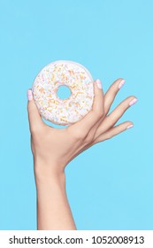 Manicure. Hand With Pastel Nails Holding Donut. Close Up Of Female With Beauty White Manicure Holding Sweet Donut In Hand On Blue Background. High Quality Image.